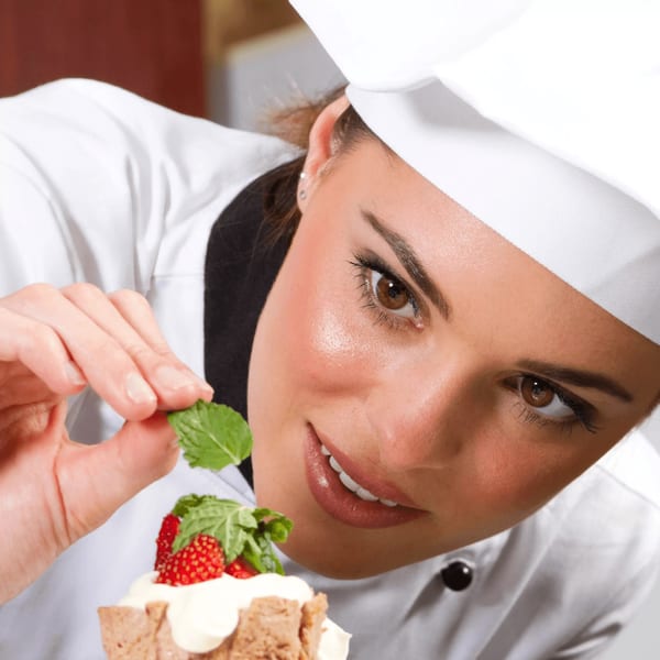 A chef in a white uniform garnishes a dessert with strawberries and mint leaves.