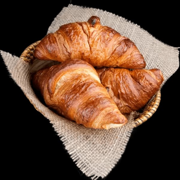 Three golden-brown croissants nestled in a woven basket lined with burlap, against a dark background.