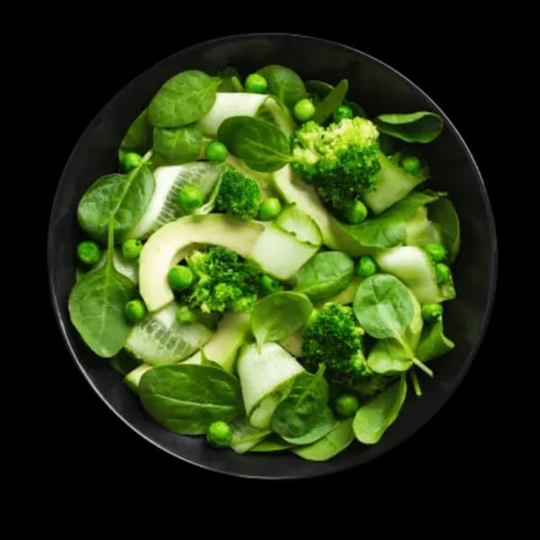 A vibrant green salad in a black bowl, featuring spinach, peas, broccoli, cucumber, and avocado.