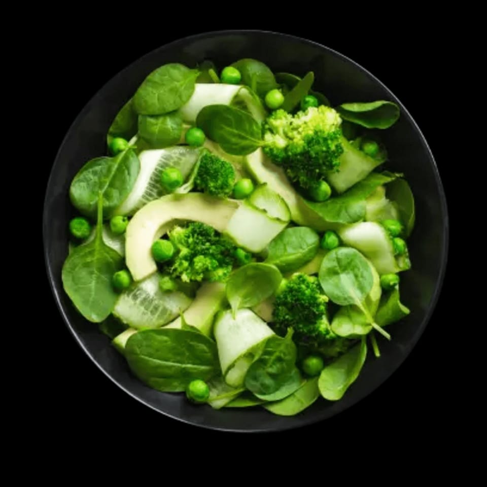 A vibrant green salad in a black bowl, featuring spinach, peas, broccoli, cucumber, and avocado.