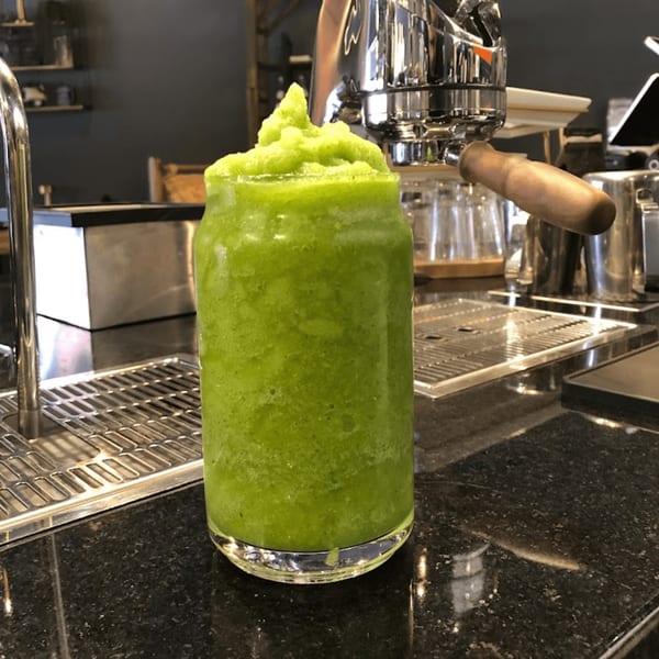 A vibrant green smoothie in a glass on a dark countertop, with espresso machine in background.