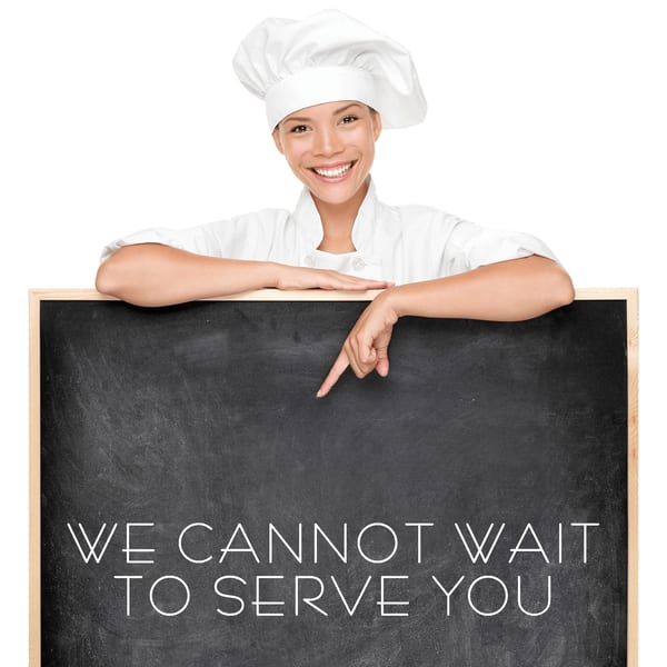 Smiling chef in white uniform and hat, pointing to a chalkboard that says "WE CANNOT WAIT TO SERVE YOU".