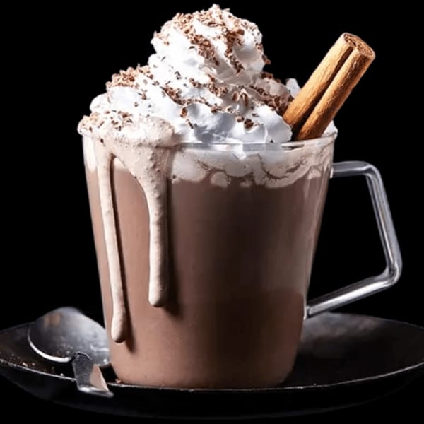 Hot chocolate in a clear mug with whipped cream, chocolate shavings, and a cinnamon stick.